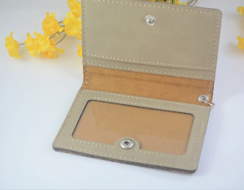 May include: A beige leather card holder with a clear plastic window for displaying an ID card. The holder has a brown suede lining and a silver snap closure.