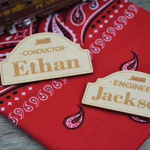 May include: Two wooden name tags with a train theme. The top tag reads "Conductor Ethan" and the bottom tag reads "Engineer Jackson". The tags are on a red bandana with a paisley pattern.