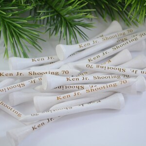 May include: A pile of white golf tees with gold lettering that reads "Ken Jr. Shooting 70".