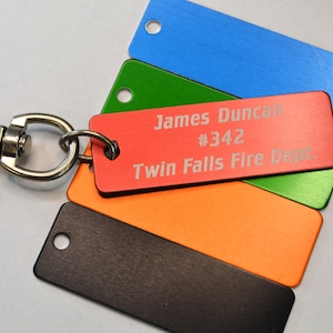 May include: A collection of rectangular keychains in various colors, including blue, green, orange, and black. One red keychain displays the text "James Duncan #342 Twin Falls Fire Dept." A silver clasp is attached to the keychains.