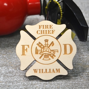 May include: A wooden firefighter's cross with the letters "FD" and the words "Fire Chief" and "William" engraved on it. The cross features a firefighter's helmet, axe, and ladder.