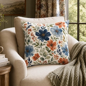 May include: A square throw pillow featuring a watercolor-style floral pattern with deep blue, light blue, and burnt orange blossoms set against a cream-colored background, accented with green leaves and small berry-like clusters on thin stems.