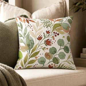 May include: A square throw pillow featuring a white background with a watercolor-style botanical print. The design includes various green leaves, stems, and small red flowers and berries scattered across the surface in a repeating pattern.