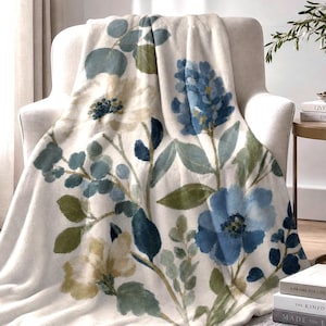 May include: A soft, cream-colored throw blanket featuring a watercolor-style botanical print. The design displays large blue and pale yellow flowers, dark blue berries, and various green leaves scattered across the fabric surface.