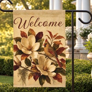 May include: A rectangular garden flag featuring the word Welcome in brown cursive script at the top. The design displays two large cream-colored magnolia flowers, dark brown and green leaves, and a cardinal bird perched on a branch.