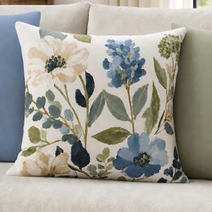 May include: A square throw pillow featuring a cream-colored background with a botanical print. The design includes large watercolor-style flowers in shades of blue, beige, and white, accented by green and dark navy leaves and stems.