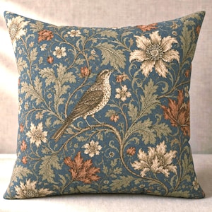May include: A square throw pillow featuring a blue background with a detailed botanical pattern of cream-colored flowers, sage green foliage, and rust-colored leaves. A brown bird is perched on a branch in the center of the design.