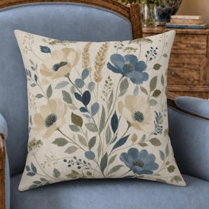 May include: A square throw pillow featuring a cream-colored fabric base with a botanical print. The design displays various flowers in shades of muted blue, beige, and sage green, interspersed with delicate stems, leaves, and wheat-like stalks.