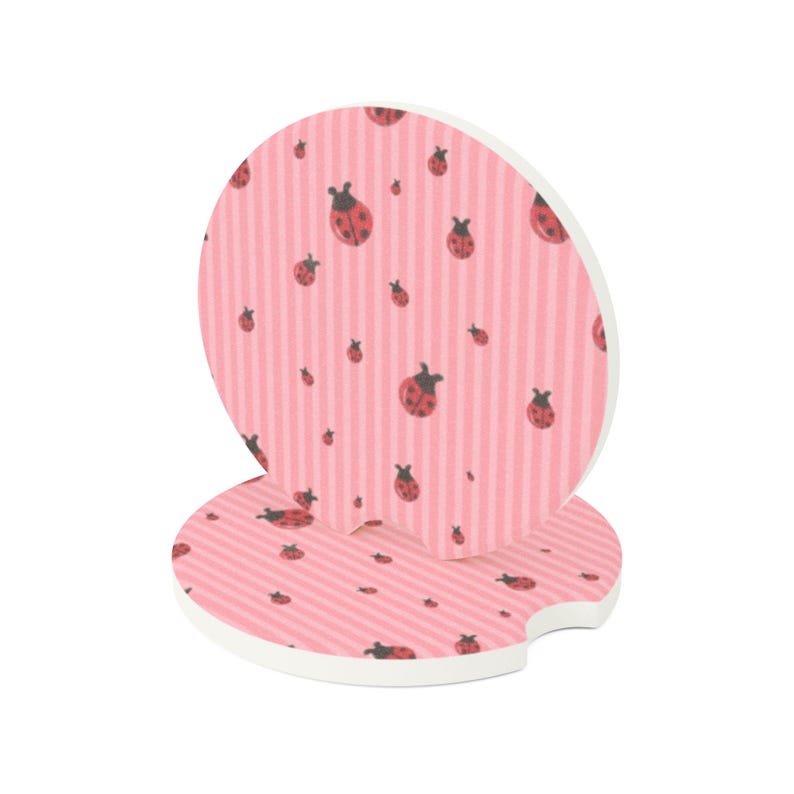 May include: Two circular car cup holder coasters featuring a light pink background with vertical stripes and a repeating pattern of small red and black ladybugs. Each coaster has a small semi-circular notch cut out from the edge of the disc.