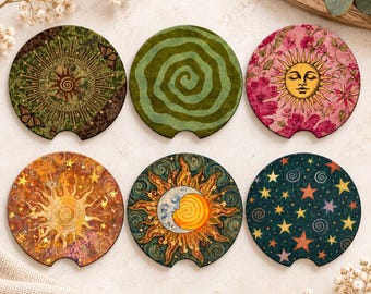 May include: A set of six circular car coasters featuring diverse designs including a sun with butterflies, a green spiral, a sun with pink flowers, a golden sun with stars, a sun and moon celestial motif, and a dark blue background with stars.