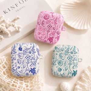 May include: Three AirPod cases with marine life patterns. One is pink with sharks, shells, and starfish. Another is white with blue sea creatures. The third is white with teal sea creatures. The cases are on a neutral surface.
