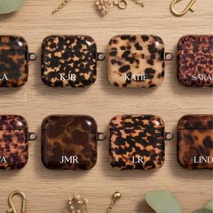May include: A collection of square-shaped protective cases for wireless earbuds, each featuring a brown and black tortoiseshell pattern. Several cases are personalized with white text including KJR, KATIE, JMR, LR, and LIND.