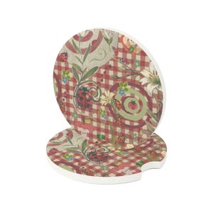 May include: Two round car coasters featuring a red and white checkered pattern decorated with ladybugs, green swirls, and white flowers. The coasters have a small notch on the side and a white edge, displaying a repeating nature-inspired print.