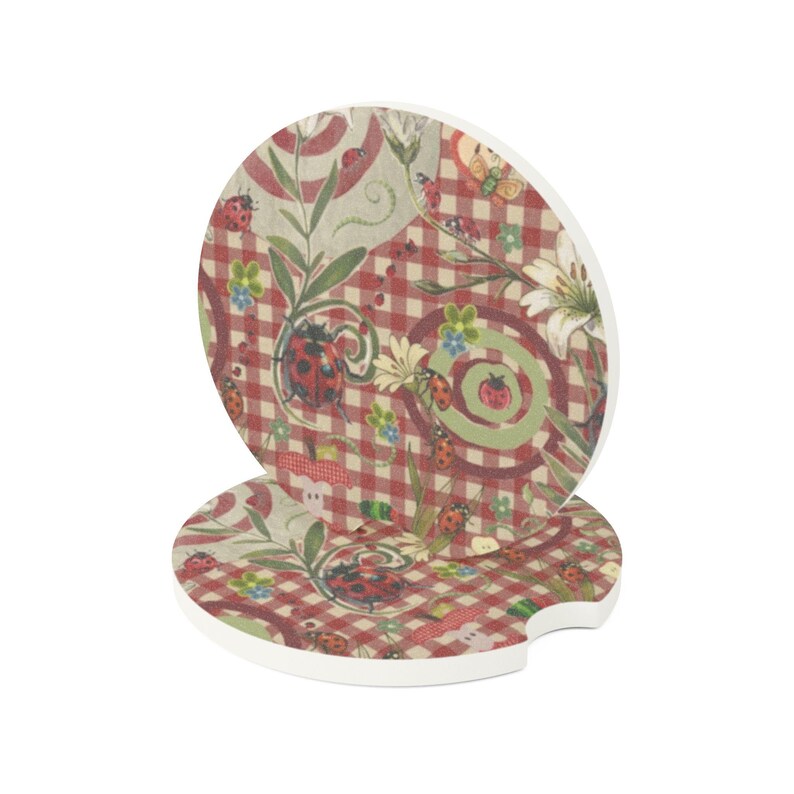 May include: Two round car coasters featuring a red and white checkered pattern decorated with ladybugs, green swirls, and white flowers. The coasters have a small notch on the side and a white edge, displaying a repeating nature-inspired print.