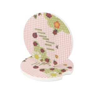May include: Two round car coasters featuring a light pink and white gingham pattern, decorated with red ladybugs, green scalloped shapes, and white daisies. The text reads: One of the greatest challenges in life is being yourself in a world that's trying to make you like everyone else.
