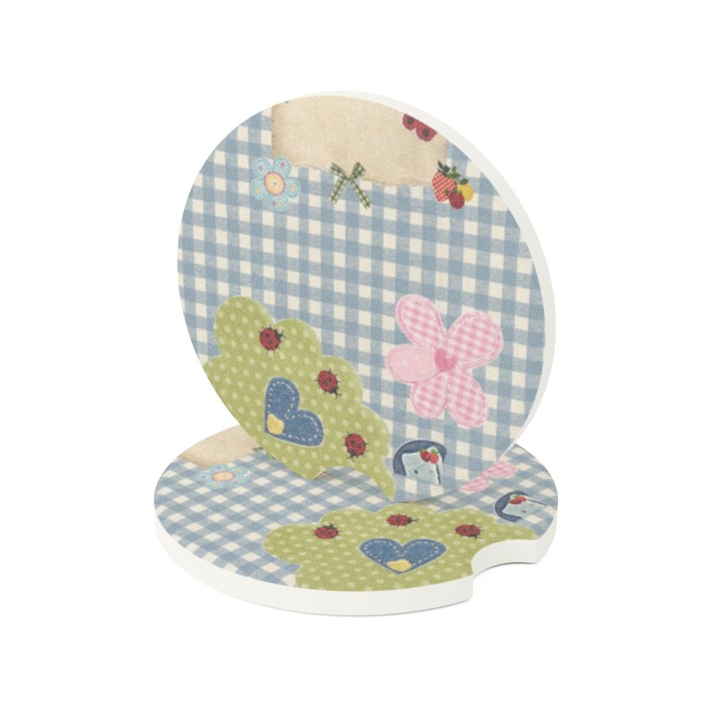 May include: Two round ceramic car coasters featuring a light blue and white gingham pattern. The design includes green cloud-shaped motifs with red ladybugs, blue hearts, pink checkered flowers, and small strawberry illustrations on a white base.