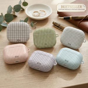 May include: Six gingham-patterned AirPod cases in various pastel colors: gray, green, light blue, pink, and lavender. Each case is personalized with the initials "RM". The cases are displayed on a wooden surface with a small dish holding rings and a pen.