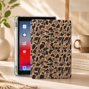 May include: A tablet case featuring a tan, brown, and black camouflage pattern with small silhouettes of flying ducks. The case is shown covering a tablet with a visible screen displaying app icons including Calendar, Clock, Maps, News, and Safari.