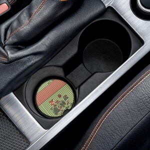 May include: A circular car cup holder coaster featuring a green and white gingham pattern, a diagonal coral-colored stripe, and a small cluster of red strawberry illustrations. The coaster is placed inside a black plastic vehicle cup holder.