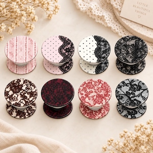 May include: Eight circular phone grips arranged in two rows, featuring various patterns including pink stripes, black lace on pink or white polka dots, solid black lace, brown floral lace, solid dark red lace, red floral lace, and black floral lace.