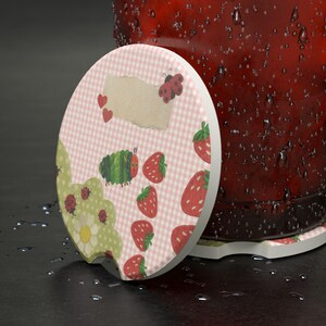 May include: A round ceramic car coaster featuring a light pink gingham pattern, a green caterpillar, red strawberries, ladybugs, and a white flower. The coaster has a small notch on the edge and is positioned next to a dark red, water-covered container.