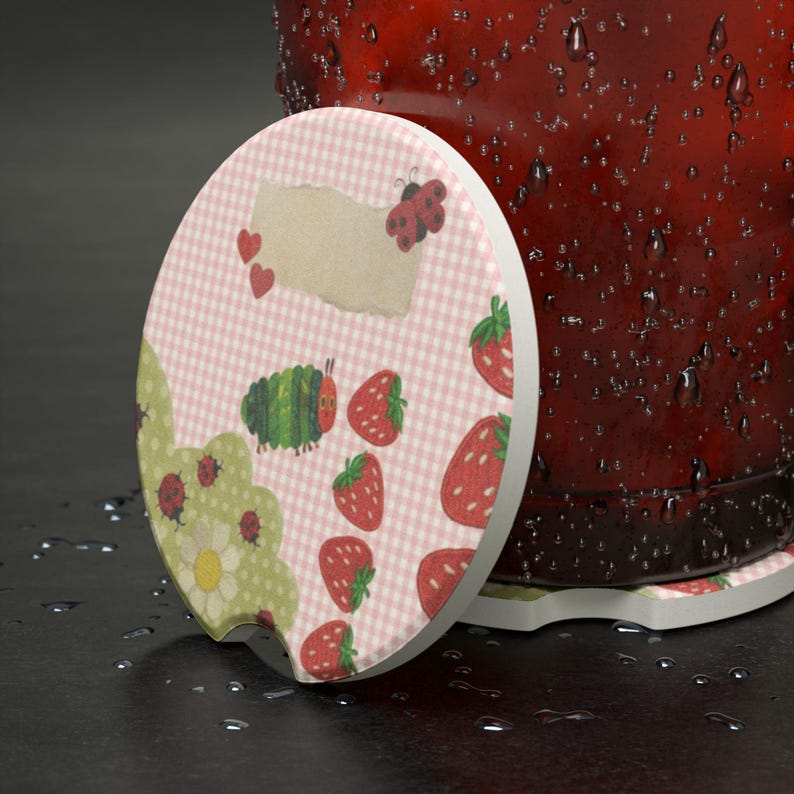 May include: A round ceramic car coaster featuring a light pink gingham pattern, a green caterpillar, red strawberries, ladybugs, and a white flower. The coaster has a small notch on the edge and is positioned next to a dark red, water-covered container.
