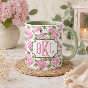May include: A ceramic mug with a sage green interior and handle, featuring a repeating pattern of pink hydrangea flowers and green leaves. A central rectangular frame with a bamboo-style border contains the pink monogram letters BKL.