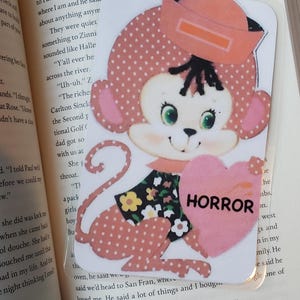 May include: A pink monkey with a red hat and a pink heart that says "HORROR" on it. The monkey is wearing a pink and black polka dot dress with yellow flowers.