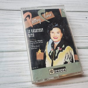 May include: A vintage cassette tape featuring Patsy Cline's 12 Greatest Hits. The cover art features a color portrait of Patsy Cline in a cowboy hat and jacket, with a gold star and the text "12 Greatest Hits" in a black and white font.