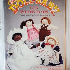 May include: A vintage sewing pattern titled "Doll Baby Easy Fashions to Sew." The cover features four dolls in various outfits, including a white dress, a pink dress, and brown overalls. The pattern includes mix-and-match outfits and full-size patterns.