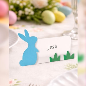 May include: A light blue bunny-shaped place card holder with the name "Josh" written on it. The card is decorated with green grass cutouts at the bottom. Easter-themed table setting with pastel eggs and flowers in the background.