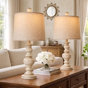 Puede incluir: Dos lámparas de mesa color crema con pantallas de tela beige sobre una mesa de madera. Las lámparas tienen una base torneada y un remate de madera. Un ramo de flores blancas y libros también están sobre la mesa.