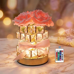 Puede incluir: Una lámpara decorativa con base dorada y diseño de cristal apilado. Cuenta con un lazo rosa y está rematada con dos rosas rosas. Un mando a distancia con opciones de color está al lado.