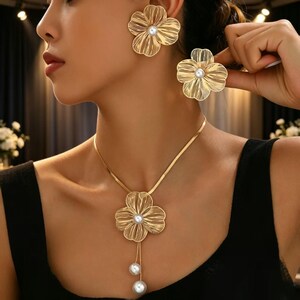 Puede incluir: Un conjunto de joyería en tono dorado que consta de un collar y pendientes a juego. Cada pieza presenta un diseño de flor texturizada de varios pétalos con una perla blanca central. El collar incluye una cadena plana y dos perlas colgantes.