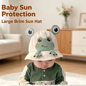 Puede incluir: Un sombrero de pescador color crema con un diseño de cara de rana verde y dos ojos saltones en la parte superior. El sombrero incluye el texto Baby Sun Protection Large Brim Sun Hat. La rana tiene una boca sonriente y pequeñas patas.