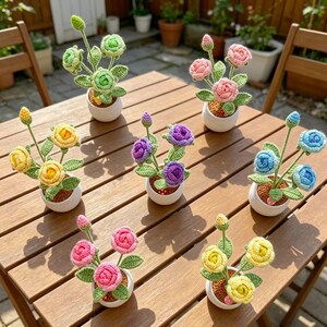 Puede incluir: Una colección de arreglos florales de ganchillo hechos a mano en pequeñas macetas blancas. Las flores son de varios colores, incluyendo verde, rosa, amarillo, morado y azul. Los arreglos se exhiben sobre una mesa de madera.
