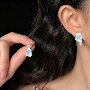 Puede incluir: Pendientes plateados con diseño de corazón y relleno de esmalte azul claro. Cada pendiente presenta una pequeña perla redonda en la parte inferior y una perla más pequeña en la parte superior. Los pendientes se muestran en una oreja y se sostienen en una mano.