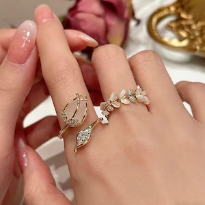 Puede incluir: Dos anillos dorados con detalles de piedras preciosas y cristales. Un anillo presenta un diseño en forma de tulipán con una piedra preciosa grande, de forma ovalada y adornos de cristal. El otro anillo tiene un diseño de hoja con pequeñas piedras preciosas.