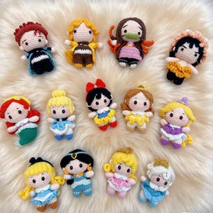 Puede incluir: Una colección de muñecas de ganchillo, cada una con características y atuendos únicos. Las muñecas tienen varios colores de cabello y visten vestidos y disfraces. Las muñecas están dispuestas sobre una superficie blanca y esponjosa.