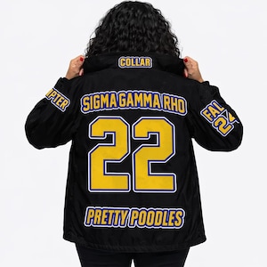 May include: A black windbreaker jacket featuring yellow and blue text and graphics. The back displays 'SIGMA GAMMA RHO', the number '22', and 'PRETTY POODLES'. The sleeves show 'CHAPTER' and 'FALL 24', with 'COLLAR' printed on the back of the neck.