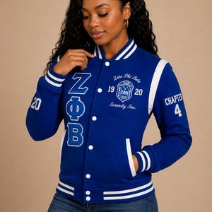 May include: A royal blue varsity jacket with white striped ribbed trim on the collar, cuffs, and hem. The front features white embroidered Greek letters, the text Zeta Phi Beta 1920 Sorority Inc., and the number 20 on the sleeve and Chapter 4.