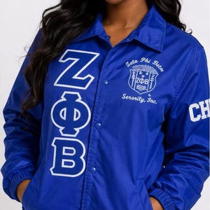 May include: A royal blue jacket with white lettering and a crest. The jacket features the Greek letters ZΦB on the front and the words "Zeta Phi Beta" and "Serority, Inc." on the chest. The jacket has a button-up front and pockets.