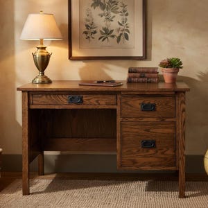 May include: A wooden desk in a warm brown finish. It has a drawer and two further drawers, all with black handles. A lamp with a beige shade and a decorative base sits on the desk, along with books and a small potted plant.