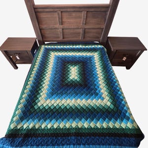 May include: A wooden bed frame with matching nightstands on either side. The bed is covered with a blue, green, and white quilted blanket. The blanket has a square pattern design. The nightstands have drawers.
