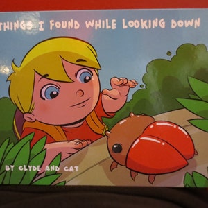 May include: A cartoon illustration of a child looking down and finding a red and brown bug. The book cover reads "Things I Found While Looking Down" by Clyde and Cat.