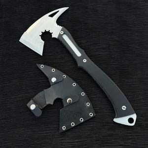May include: A tactical axe with a silver blade and a black handle. The axe head features a spike and a gear-shaped cutout. A black leather sheath is included to protect the blade. The axe is designed for outdoor use.