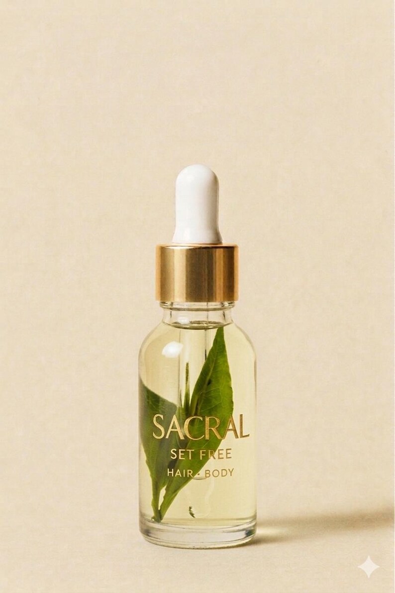 May include: A clear glass bottle of Sacral Set Free Hair Body oil with a white dropper and gold accents. The bottle contains a green leaf and the text "SACRAL" in gold lettering. The background is a light beige color.
