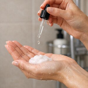 May include: A close-up of hands holding a dropper dispensing a clear liquid onto a dollop of white foam. The dropper has a black top and a clear glass tube. The background is a blurred bathroom setting.