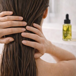 May include: A person's wet, brown hair is held with both hands. A bottle of Sacral Set Free hair and body oil is in the background. The bottle has a black dropper top and a yellow liquid inside.