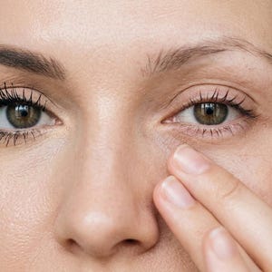 May include: Close-up of a person's face, focusing on the eyes. One eye has mascara applied, while the other eye is without. A hand gently touches the skin under the eye. The skin tone is fair.